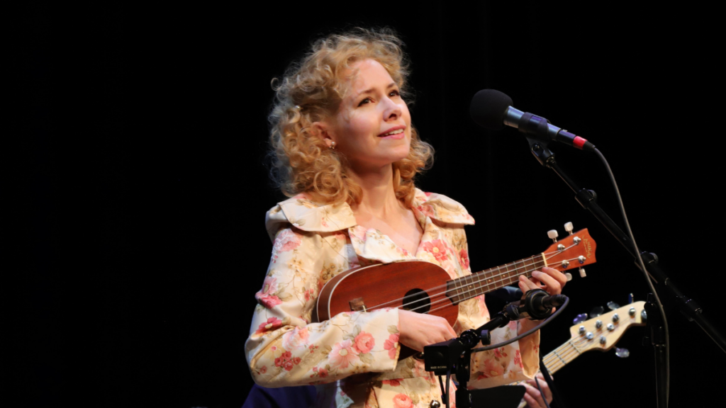 A woman with blond curly hair plays a ukelele.
