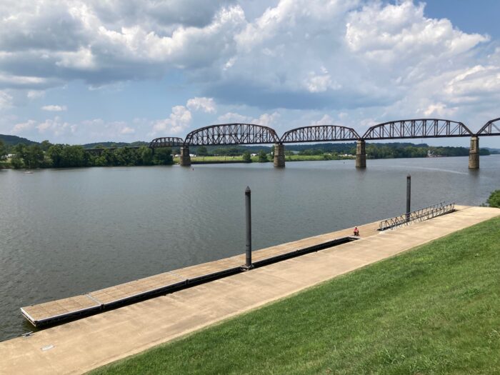 a wide river flows under a steel railroad bridge and a sunny sky with white clouds.