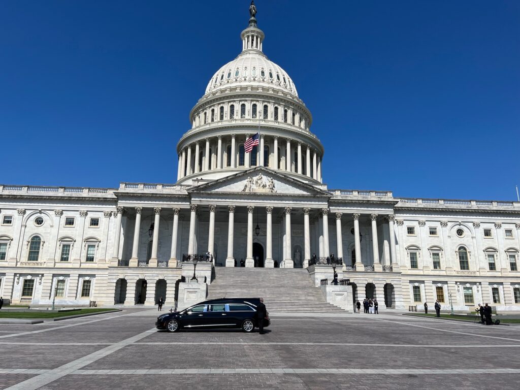 A black hearse pulls up in front of the white steps of the U.S. Capitol against a clear blue sky on a summer day.