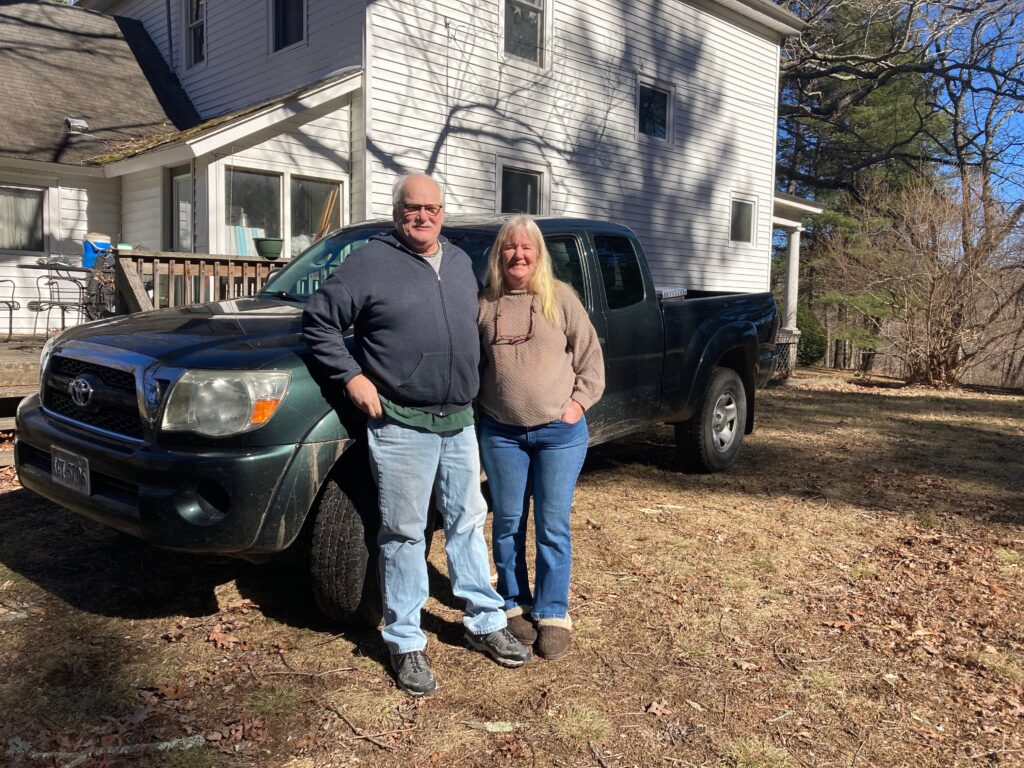 Two older people stand in front of a truck outside their home. They both are wearing jeans and long-sleeved shirts. Behind them is a white house.
