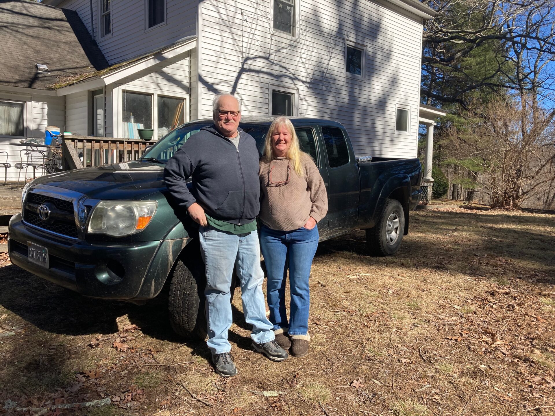 Two older people stand in front of a truck outside their home. They both are wearing jeans and long-sleeved shirts. Behind them is a white house.
