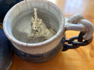 A handmade mug is shown with a braided handle. Inside the mug is a small unicorn.