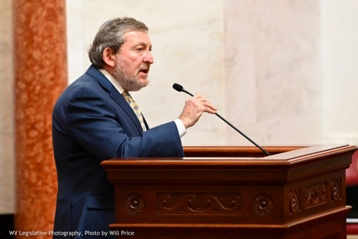 An older man, 60s, stands at a podium in the West Virginia Senate chamber. He reaches for the microphone to adjust the height. He has salt and pepper hair and wears a navy blue suit with white button shirt and tie.