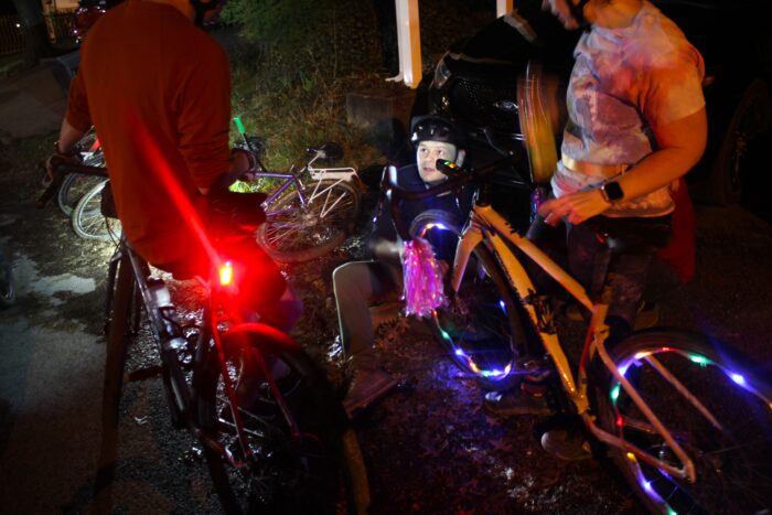 A woman stands next to her bicycle, her head cut off by the photograph's frame, as a man works on the front wheel of her bicycle. There are multicolored lights woven through the spokes of the bike's wheels. The scene is dark and lit by small lights. Another person with their back to the frame straddles a bicycle to the left of them with a red safety light illuminated.