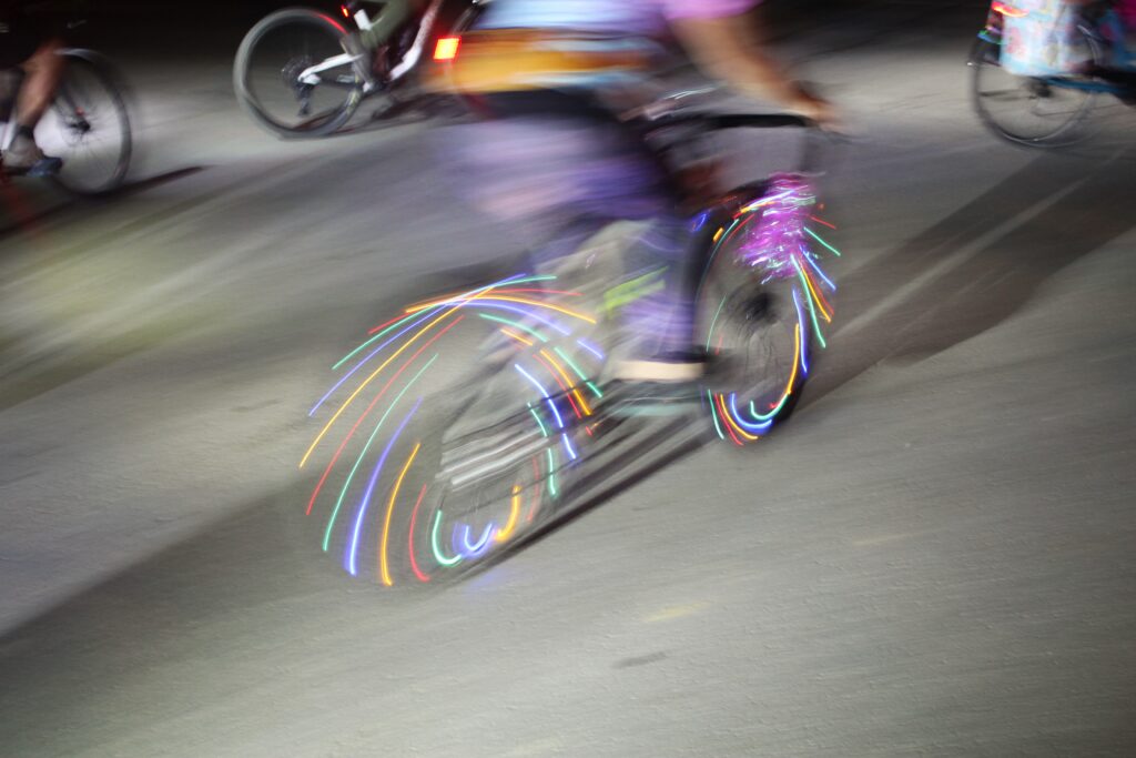 A cyclist standing up on her pedals is blurred by motion. The bicycle she rides has multicolored lights woven through the spokes of the wheels. The image is lit from the left by a bright white light, casting long shadows to the right of frame. In the background more bicycles, less blurred by motion, can be seen.