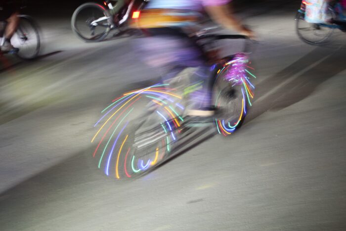 A cyclist standing up on her pedals is blurred by motion. The bicycle she rides has multicolored lights woven through the spokes of the wheels. The image is lit from the left by a bright white light, casting long shadows to the right of frame. In the background more bicycles, less blurred by motion, can be seen.