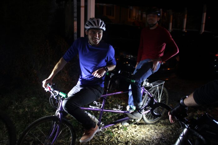Two men sit on a purple tandem bike in the dark, lit by small lights from the left and front. The man in the front wears a blue shirt with the sleeves pushed up, while the one in the back wears an orange-red shirt. Both are wearing jeans and helmets.