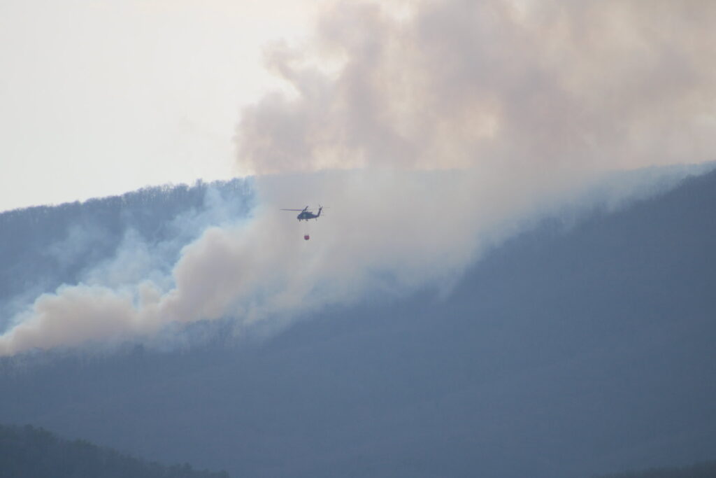 Smoke rises off of a mountain as a National Guard helicopter dispenses water over wildfires down below.