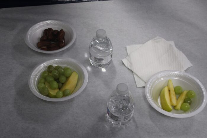 Styrofoam bowls and small bottles of water are set on a white paper sheet with white square napkins. The bowl in the top left of frame holds dates, and the bowl below that contains green grapes and apple slices. In the center of frame stand the two small plastic water bottles, and in the bottom right corner is another bowl of apple slices and green grapes with a few napkins above.