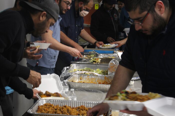 A row of aluminum trays filled with food pushes out into the frame, with men congregated around the trays. The men are serving themselves the food, which includes seasoned rice and salad.