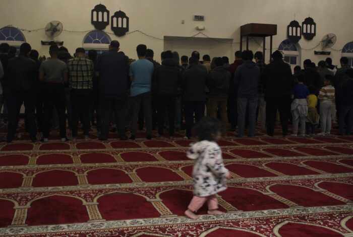Men facing away from the camera stand on a red carpet with a pattern of filligreed arches. In the foreground a small girl wearing a white jacket and pink pants can be seen running across the frame.