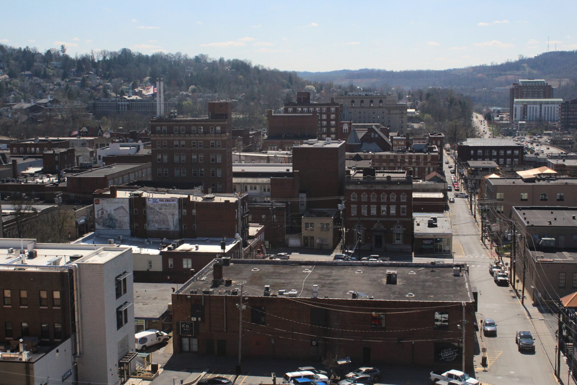 A view of downtown Morgantown is taken from inside of the West Virginia University astronomical observatory. Streets can be seen stretching away into frame around squat brick buildings. In the top right of frame can be seen a hint of the Monongahela River with the Morgantown Marriot tower in front. A white brick building with "Hotel Morgan" lettering can be seen in the middle background. A wooded ridge rises in the background, with houses dotting the hillside that slopes into downtown visible in the left background.