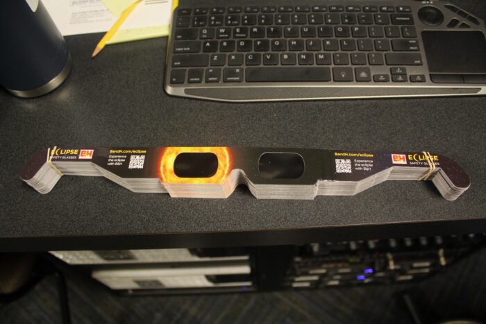 A stack of eclipse glasses, stylized with a sun over the right eye (left as oriented to the camera) are set under a keyboard on a black dress. A server stack can be seen further under the desk, as well as a pencil and papers on the desk.