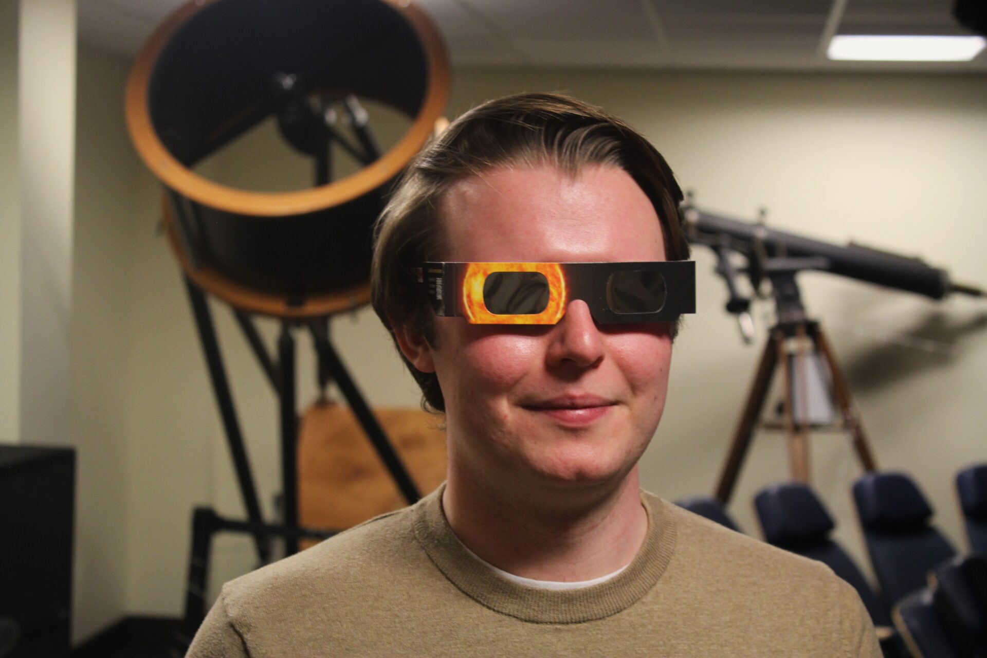 A man in a tan shirt wears eclipse glasses while standing in front of telescopes in the West Virginia University Planetarium.