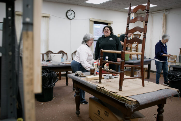 Two women, one older and one younger, talk to each other and laugh. Around them are caned chairs waiting to be restored.