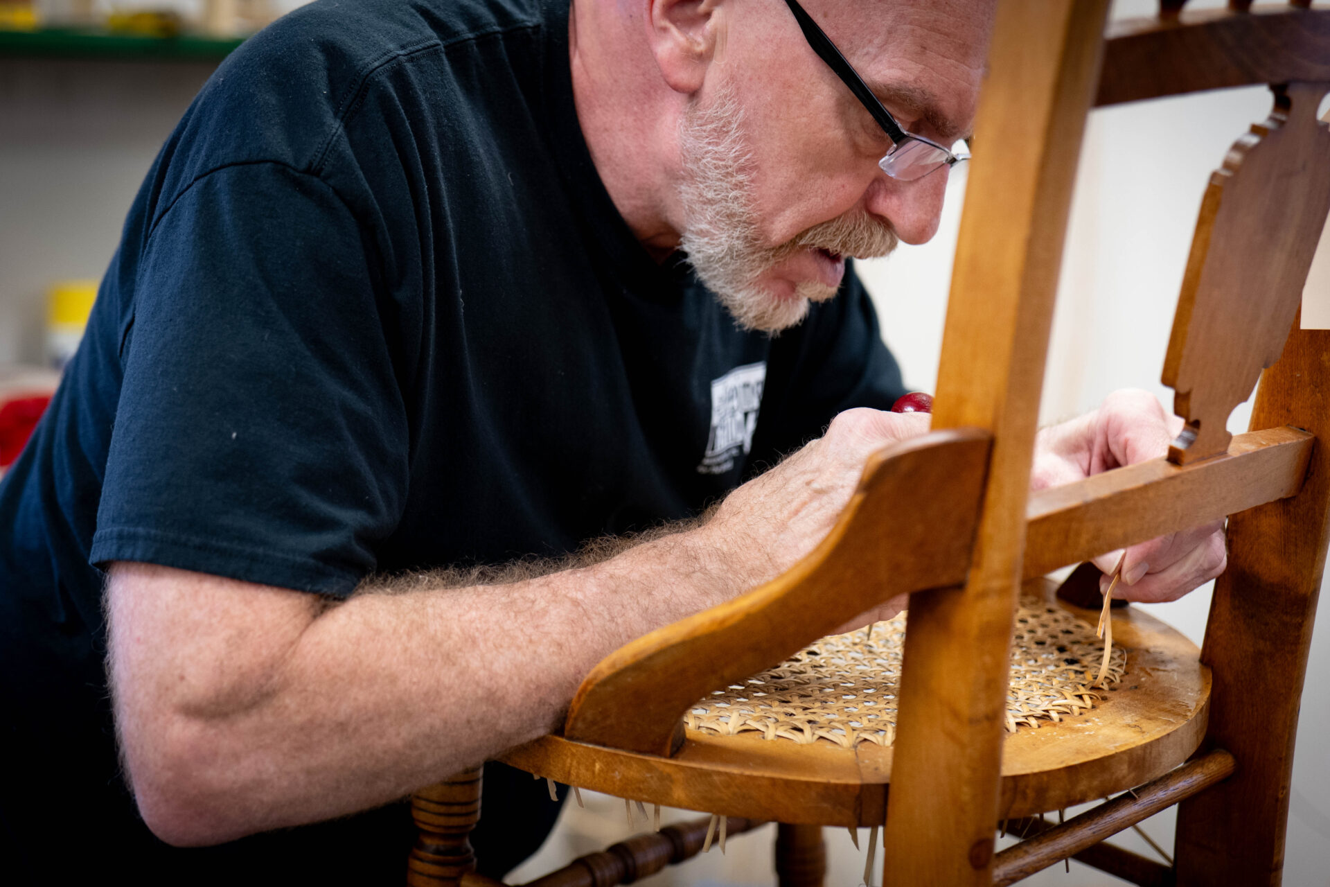 An older man wearing black glasses and with a trimmed white beard bends in close to the seat of a chair. It is a caned chair he's restoring.