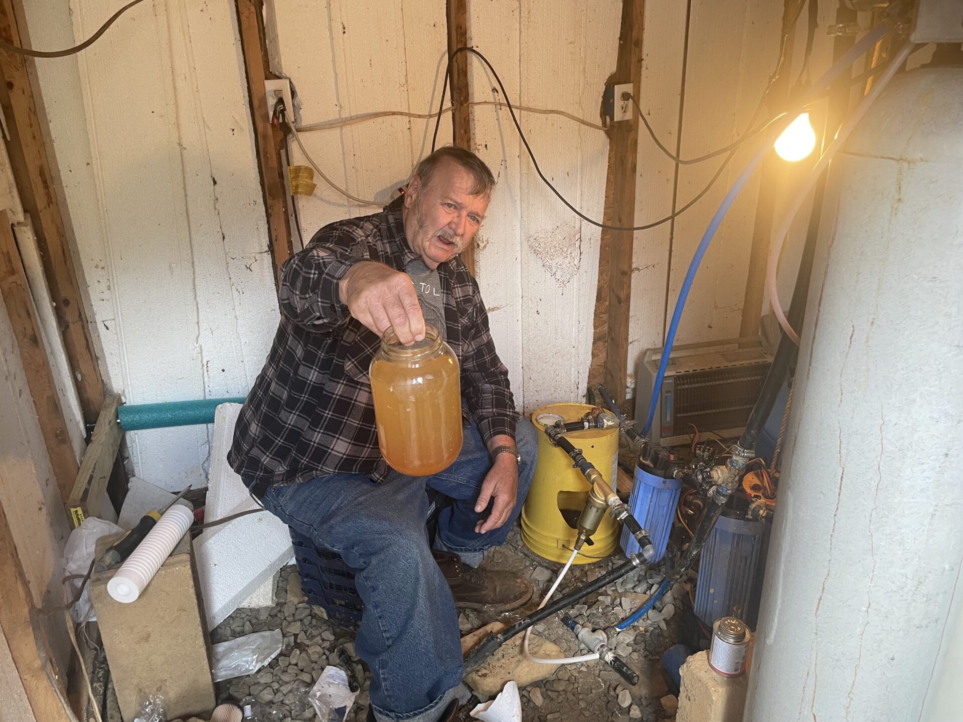 A man sits in a shed holding a mason jar of yellow foggy water. The man is older, with blue eyes, he looks upset.
