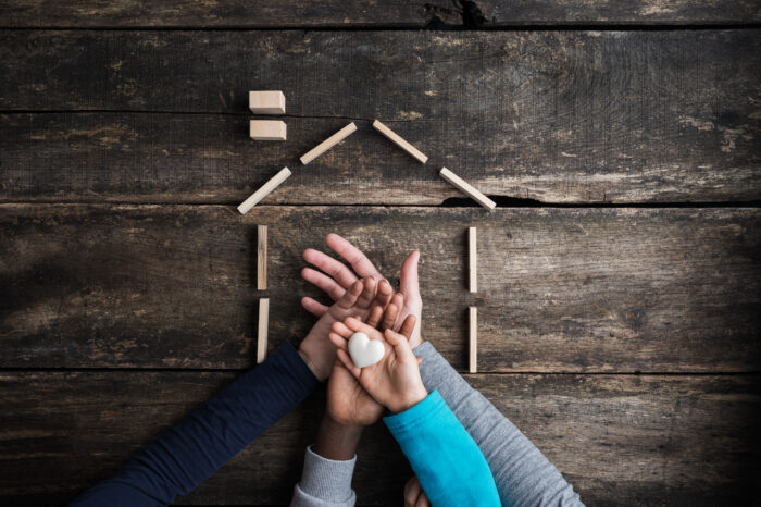 Top view of a family of four placing hands one on top of the other in a conceptual image of love, togetherness and safety. The hands are placed inside of the outline of a home made from small wooden blocks, and all is placed on a dark wood background.