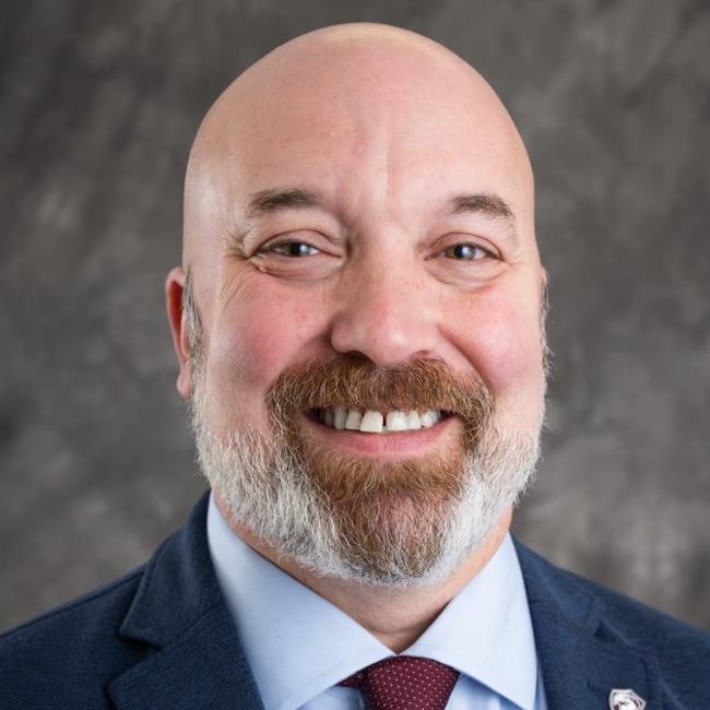 A front facing portrait of a man. He is bald with a two-tone beard of a brown goatee and grey beard elsewhere. He wears a red tie and white shirt with a blue jacket.