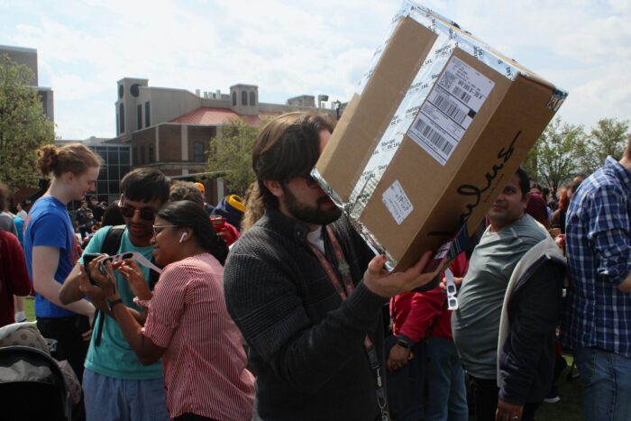A bearded man wearing a dark green sweater stands in a crowd holding a cardboard box to his eye. The box is taped closed with metallic tape and a shipping label is visible in the top right corner of the box. Behind him, people look at a phone while holding paper eclipse glasses. Buildings can be seen rising behind the man.