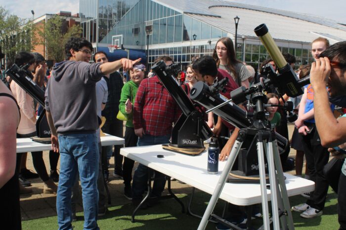 A man wearing jeans and a grey hoodie in the left of frame points across the frame of the image. In front of him and in center frame are several telescopes, black cylinders angled up towards the left of frame. Some of the telescopes are placed on white folding tables, and the two telescopes closest to right of frame are on their own metallic tripods. The rightmost and smallest telescope is gold in color. Men are positioned to use two of the telescopes, the leftmost on the table and the rightmost golden telescope.