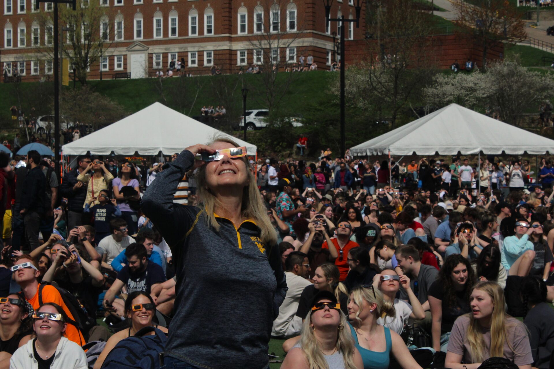 A blonde woman wearing eclipse glasses and smiling stands amidst a crowd sitting on a green. She wears a grey quarter zip sweater with a "flying WV" logo visible on the right of the sweater's chest. The crowd spreads behind her until a grassy hill rises towards a red brick building.