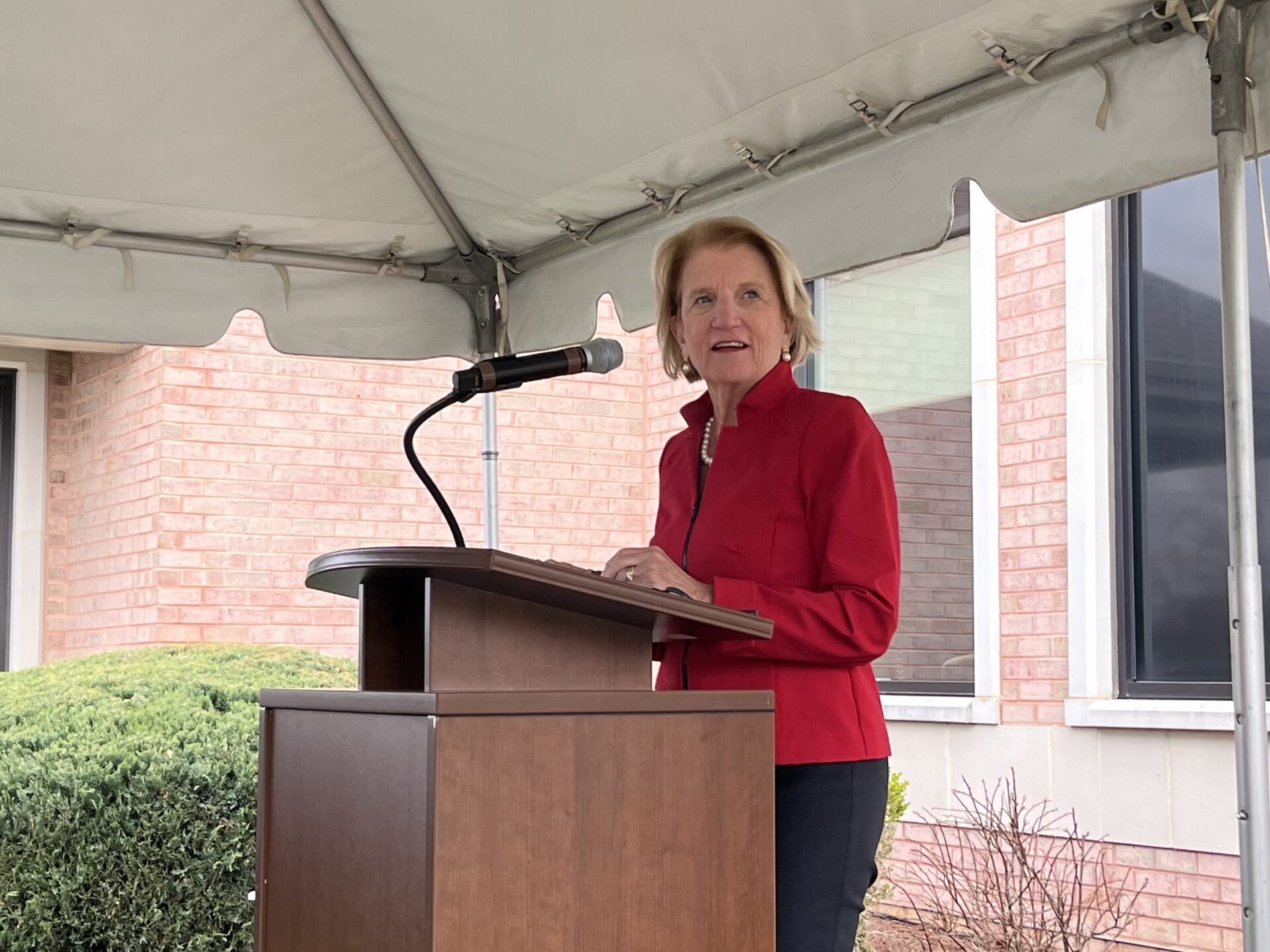 Senator Shelley Moore Capito stands at a podium, speaking into a microphone. She is wearing a blazer, and looking to her left, toward the camera.