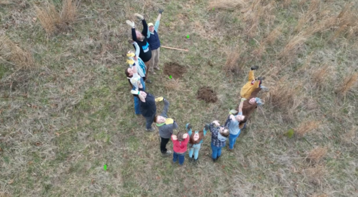 People wearing gloves stand in a half-circle with their hands in the air. Two piles of dirt are beside them, where saplings are now planted. The picture is taken by a drone from above.