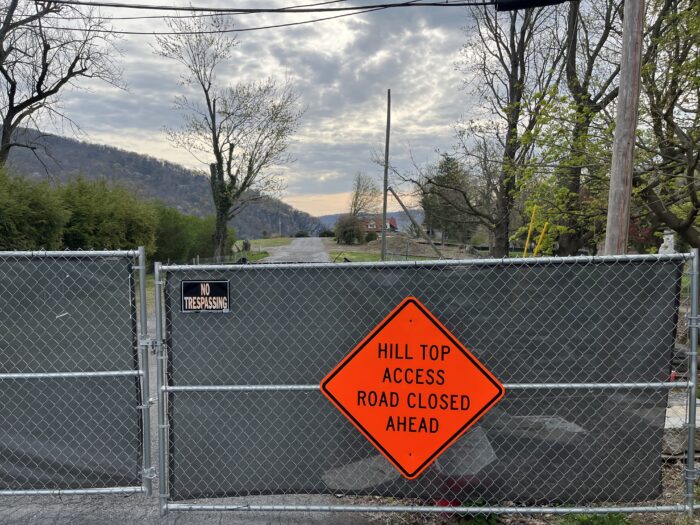 A clearing with mountains in the background is partially visible behind a chain-link fence. The fence displays signs that say "No Trespassing" and "Hill Top Access Road Closed Ahead."