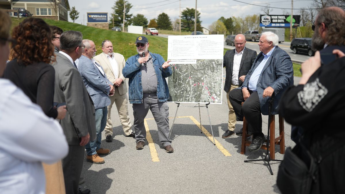 People stand around a board with a map that shows a highway and several points marked along the route in red. A man stands before the board, gesturing at it and speaking into a microphone. Gov. Jim Justice sits to the side of the board on a wooden stool. Cars and road signs are visible in the background, as the gathering is being held in a parking lot.