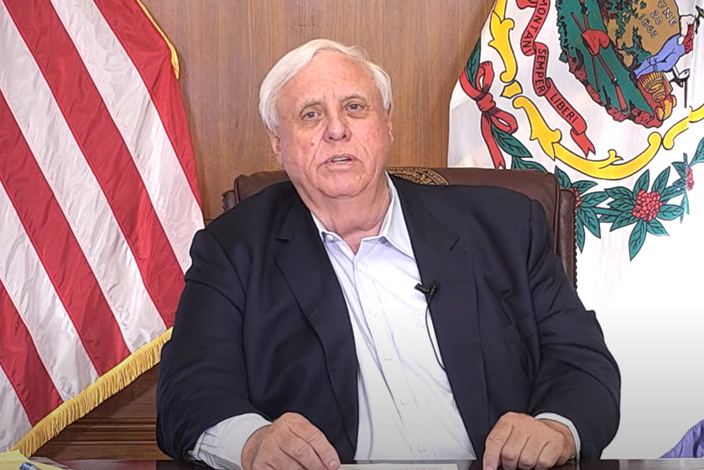 Governor Jim Justice sits at his hardwood desk, which has a piece of paper on top of it. He is wearing a suit and looking forward. Behind him stand the American and West Virginia flags.