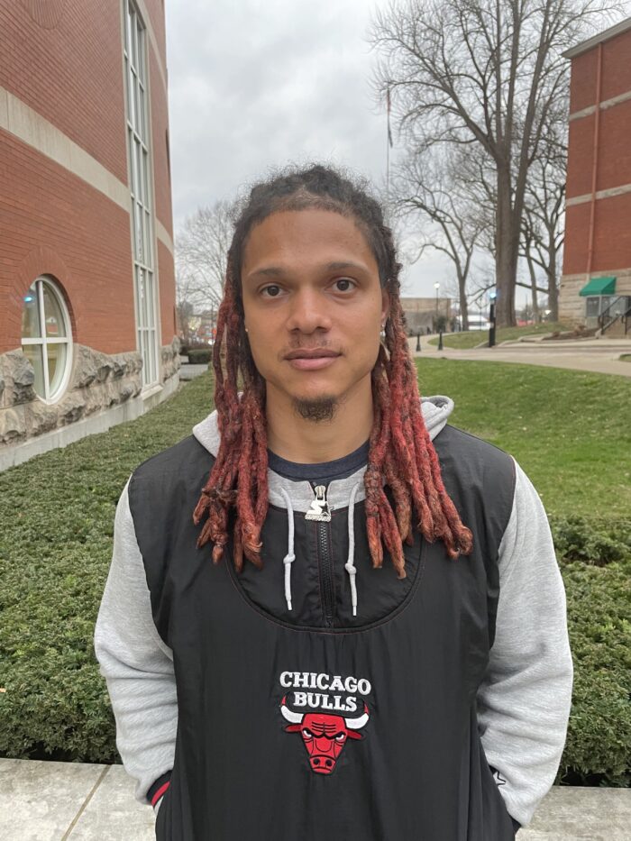 A young adult man with red and brown dreads. He wears a black and gray hoodie with the Chicago Bulls logo and mascot on the front. His hands are in his pockets.