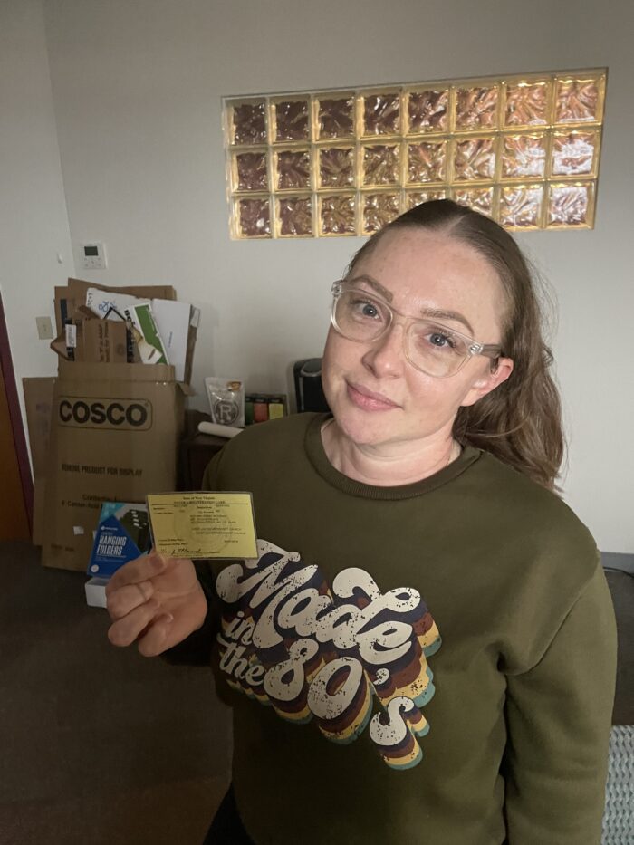 A young adult woman with brown hair pulled back into a ponytail and glasses. She holds up a yellow card for the camera and tilts her head to the side slightly. She wears an olive green sweatshirt that reads, "Made in the 80s."