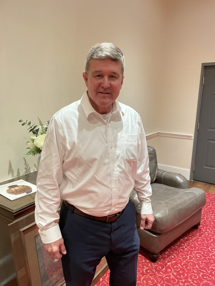 An older man with white, gray hair poses for a photo. He smiles slightly. He wears a white button up shirt and navy slacks with brown belt. Behind him is a gray chair and a bouquet of flowers in a vase on a table.