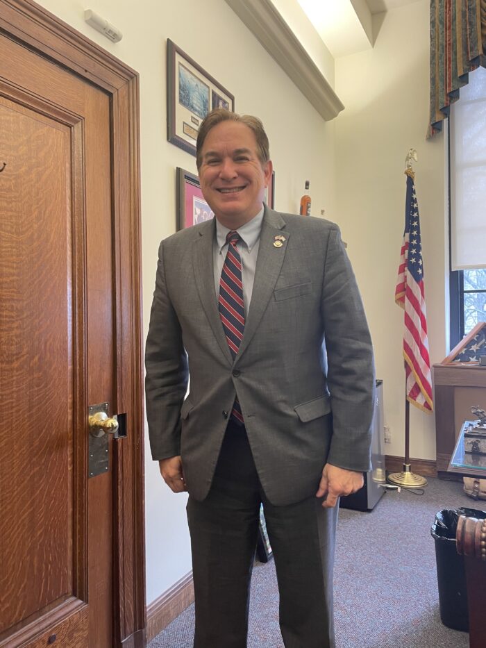A middle aged adult man smiles for a photo. He wears a gray suit and slacks, a red and blue tie, and he has a pin on his lapel. Behind him, there is an American flag and a room that looks like an office.