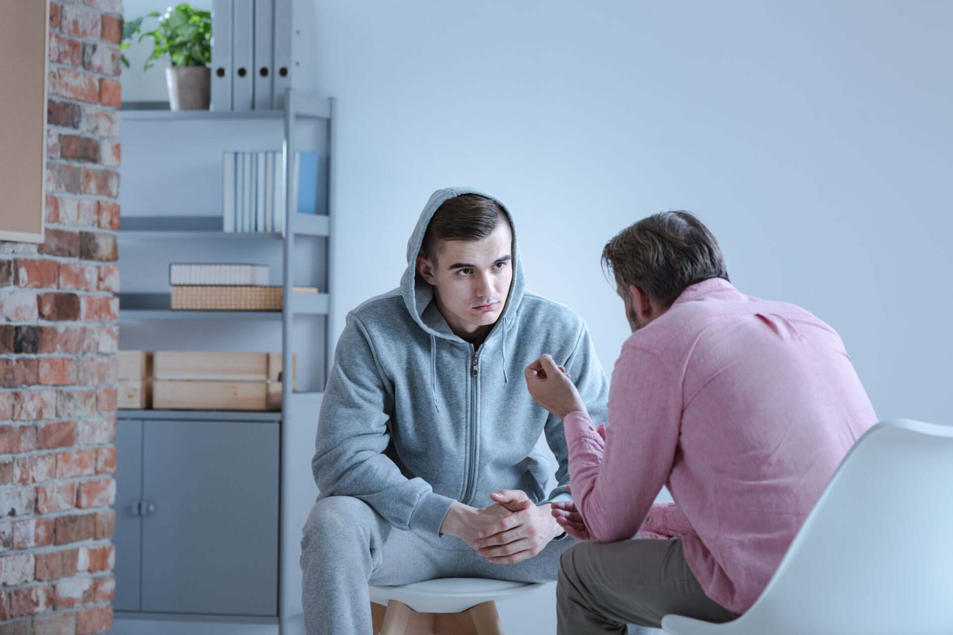 Two men are seen sitting in a room talking to one another. One wears a grey sweatsuit with the hood up around his head while the other, with his back to the camera, wears a pink shirt and appears to be encouraging the man in grey.