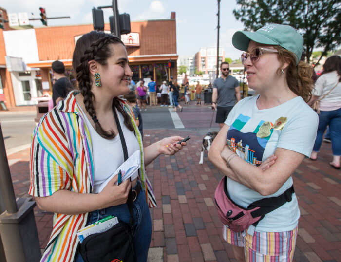 Two young women stand side by side speaking to each other. One woman holds a microphone up to the other. The woman holding the microphone has dark brown hair with braids and wears a rainbow striped button up shirt. The woman she speaks with wears sunglasses and a ball cap, t-shirt, shorts and a pink fanny pack. 
