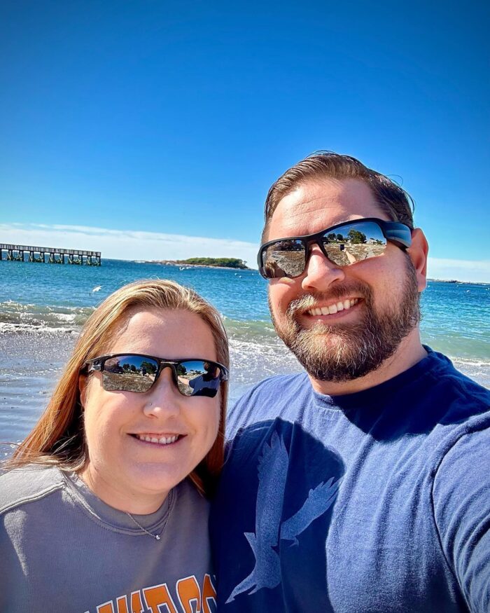 An adult woman and man pose for a selfie on the beach. The sky above them is blue with some clouds far off in the distance. Behind them is blue ocean. Both the man and woman wear sunglasses and are smiling.