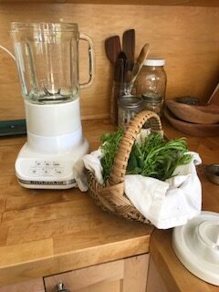 A blender is seen on a wooden countertop. Next to it is a basket full of green herbs.