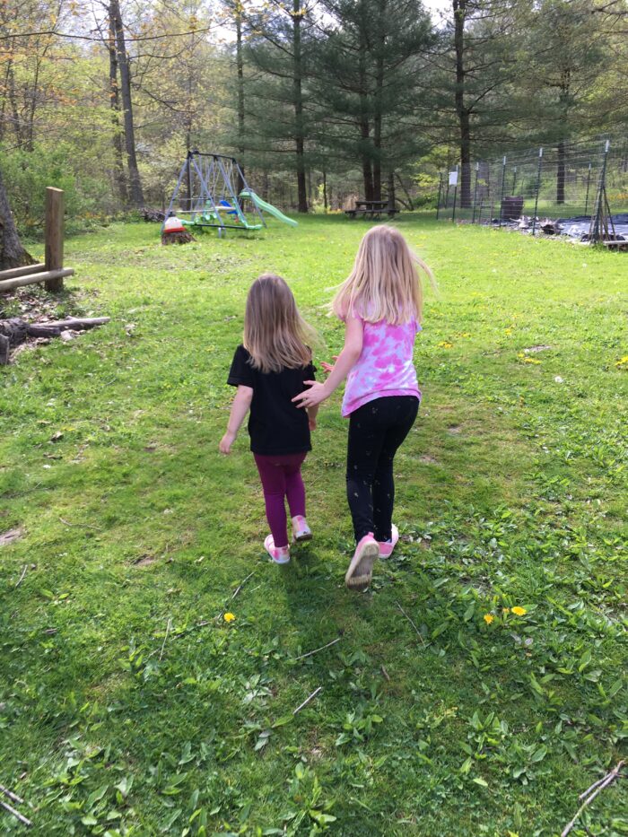Two little girls with blonde hair walk away from the camera. One girl is slightly taller than the other. One girl wears a pink and white t-shirt while the other, shorter girl, wears a black t-shirt and maroon pants. Both are wearing sneakers. They are walking in a yard toward a swing set.