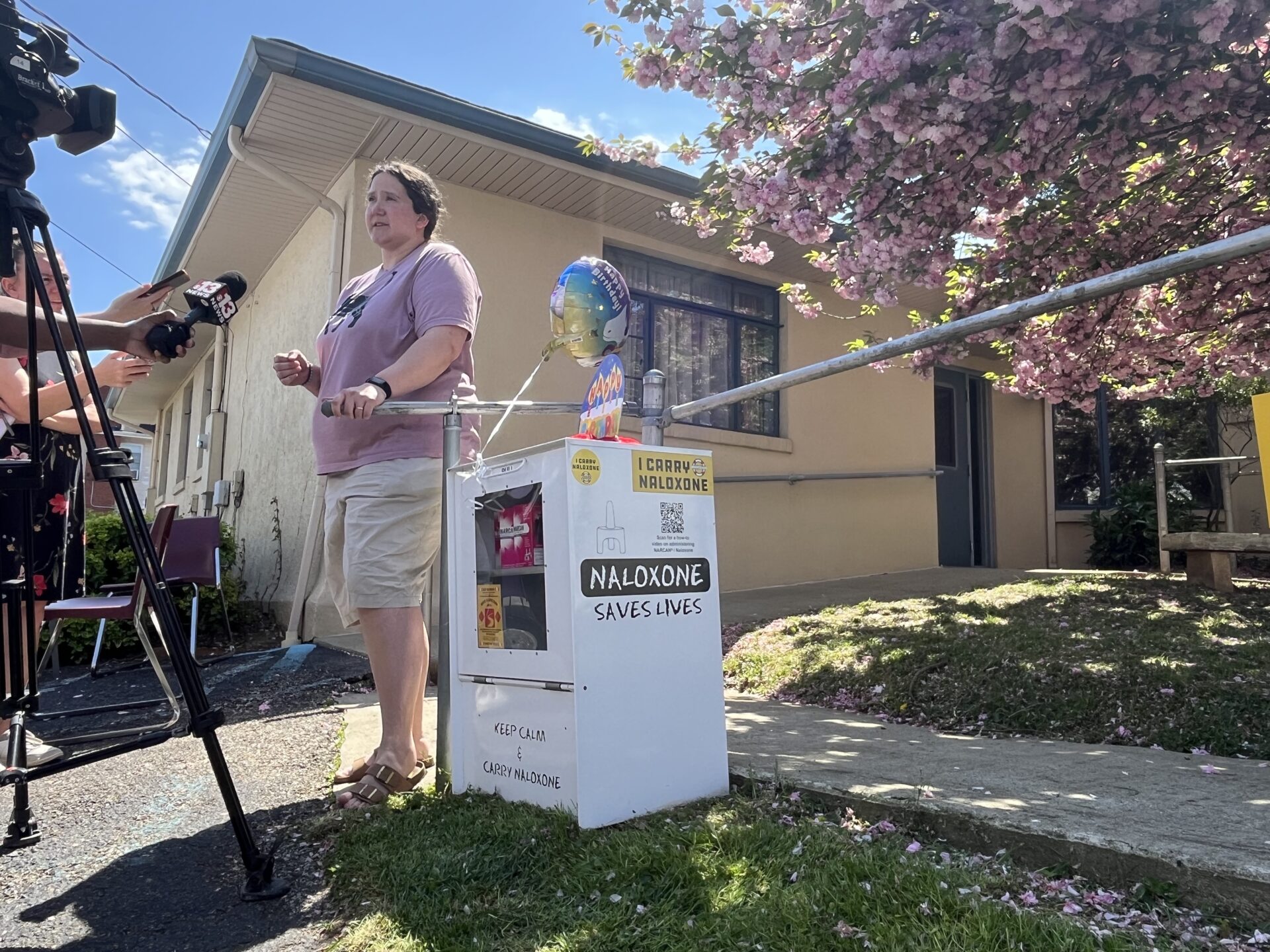A woman wearing a pink shirt and tan shorts stands next to a newspaper box containing narcan. The box is white with a window out the front. The woman is surrounded by media interviewing her about the box.