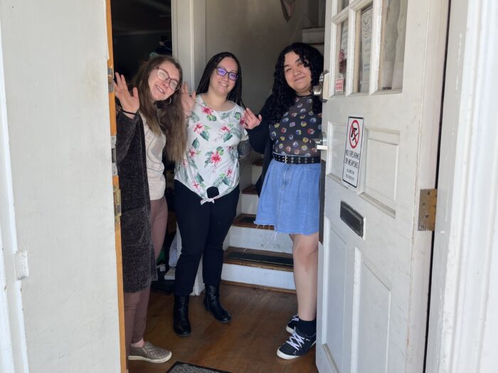 Three people stand beside a front door, waving at the entryway. The person on the far right is holding the door slightly ajar, so you can see inside.