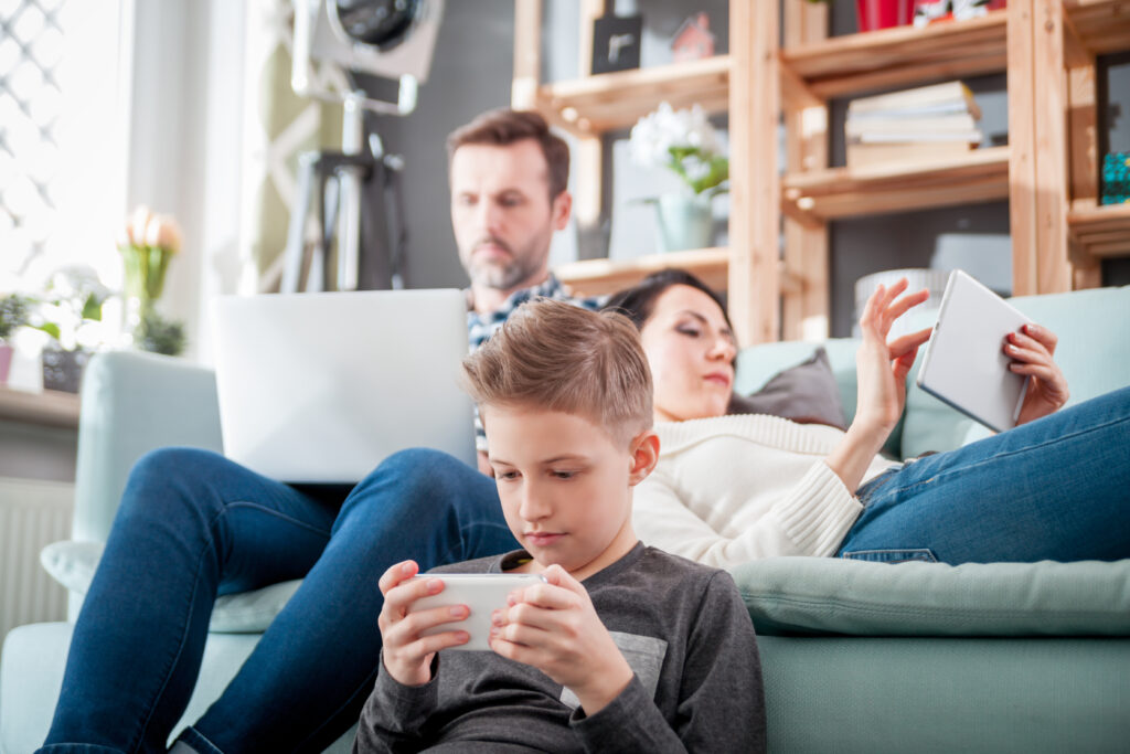 A family sits together while using different digital devices. In the foreground, a young boy looks at a smartphone while sitting on the floor in front of the couch where his parents sit. In the midground can be seen a man wearing blue jeans sitting on the left side of a couch using a laptop. To his right, a woman lays down on the couch and leans her head on the man's side while interacting with a small tablet. In the background can be seen shelving made of light wood with books and decorations.