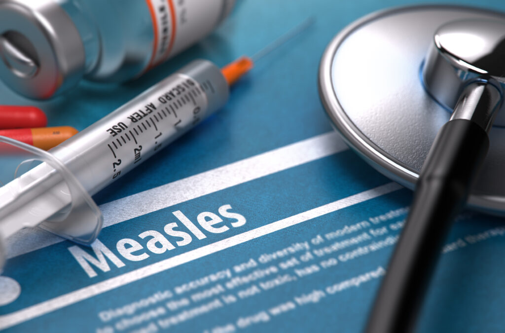 A syringe and stethoscope rest on a blue paper that reads "Measles" with facts about the virus listed below.