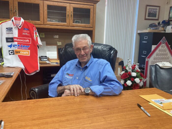 An elderly man sits at his desk with one hand on the other. He has white hair, wears glasses, and wears a blue button up shirt. Behind him is a NASCAR shirt.