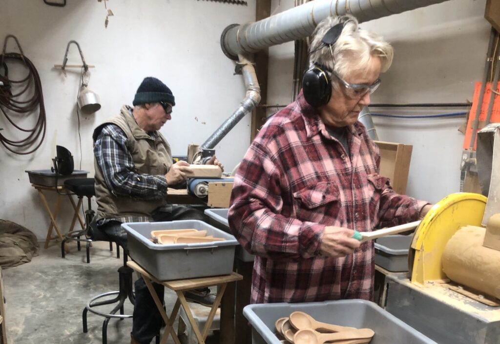 Two older people, a man and a woman, work on handmade wooden spoons in a workshop. Both people are wearing flannel shirts.