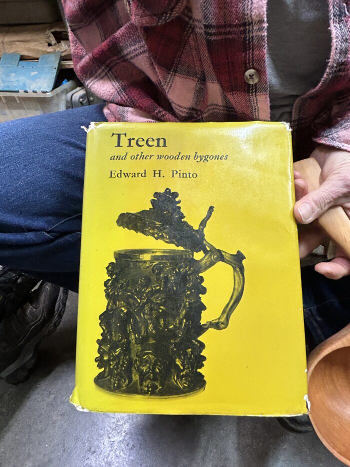 A book is being held up by a hand. The book's cover is yellow and features a black and white photograph of a mug. The title reads, "Treen and other wooden bygones" by Edward H. Pinto.