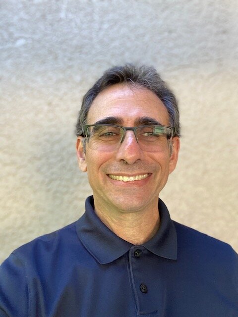 An adult, middle age man with salt and pepper hair smiles for the camera. He wears glasses and a blue polo shirt.