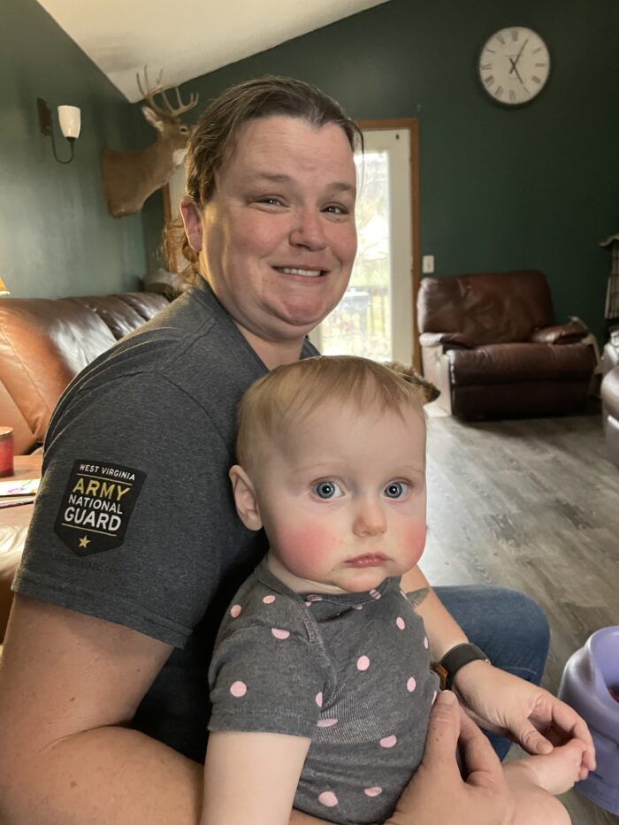 A middle age woman smiles for the camera while holding a baby in one arm. The baby looks up at the camera. The woman wears a gray t-shirt, and there's a patch on the sleeve that reads "Army National Guard." The baby has big, blue eyes, blond hair, and wears a gray shirt with pink polka dots.