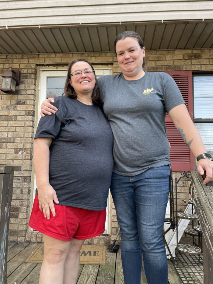 Two women stand on a porch. One woman has her arm around the other. One woman wears glasses, a gray shirt and red shorts. The other woman wears a gray shirt and jeans. Both women have dark brown hair and smile for the camera.